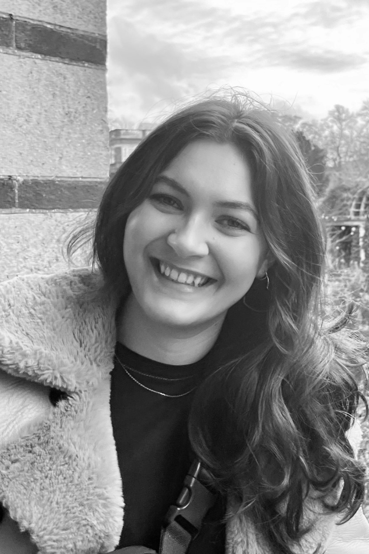A black and white image of a woman smiling at the camera. Even though the image is black and white, she has long dark hair and is wearing a fluffy jacket.